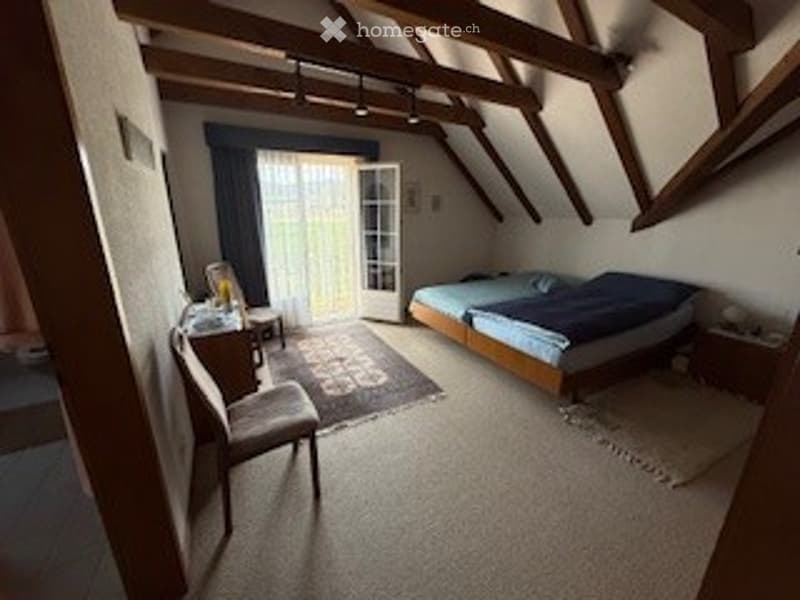 Cozy attic bedroom with wooden beams, large window with balcony access, double bed, nightstand, chair, small table, and area rug.