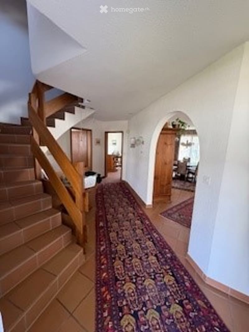 A wide hallway with a patterned carpet, wooden staircase on the left, and several doors leading to other rooms. The walls are white, and the ceiling is high with recessed lighting.