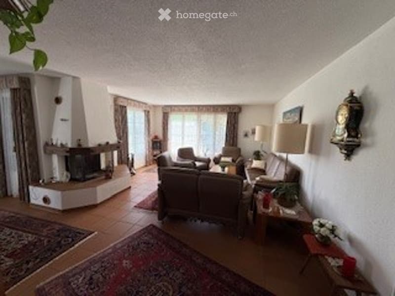 Spacious living room with a fireplace, large windows, and comfortable seating including a sofa and armchair. The room features a coffee table, side tables, and a floor lamp, creating a warm and inviting atmosphere.