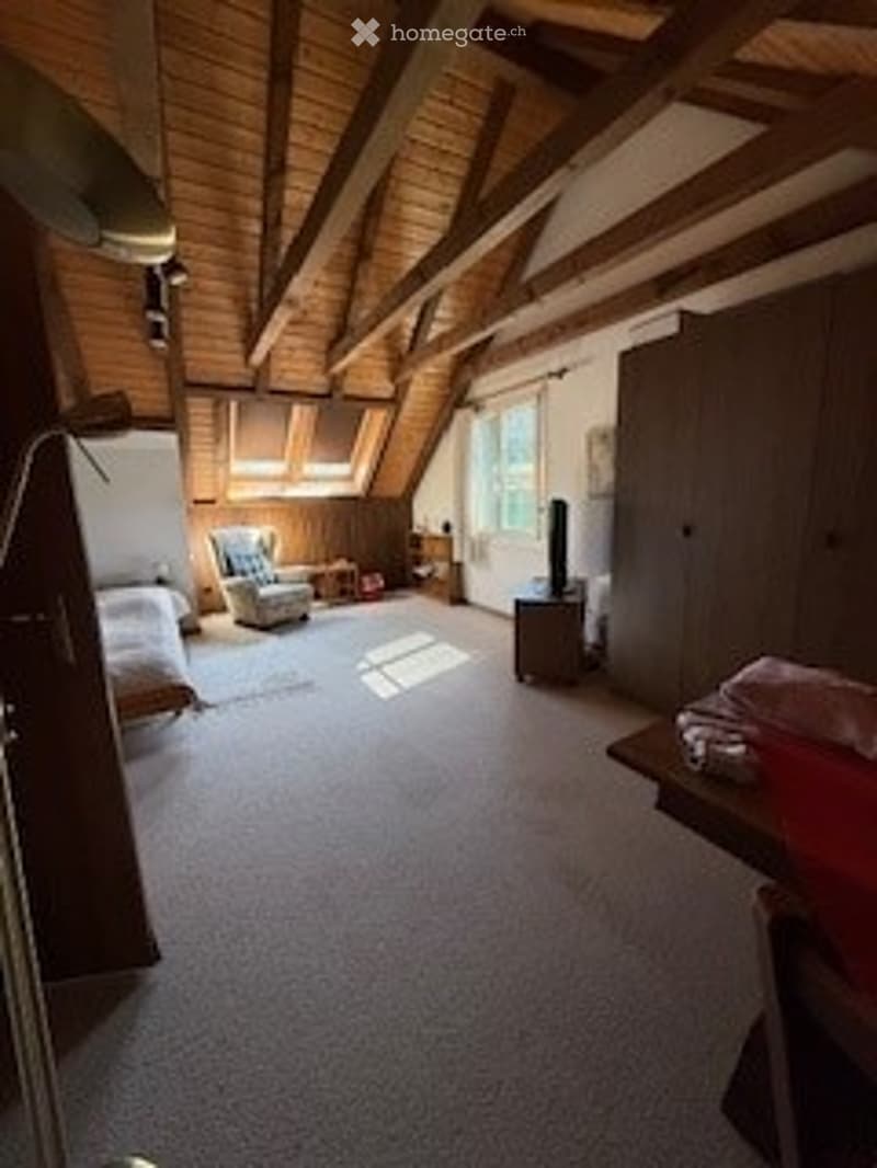 Bright attic bedroom with wooden beams, large windows, fireplace, bed, sofa, and small table.