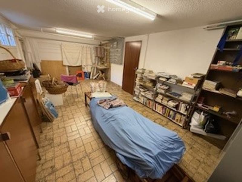 A small, multi-purpose room with a bed covered in a blue blanket, a bookshelf filled with books, a small table, and a chair. The room has tiled flooring and white walls.