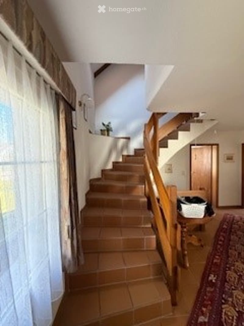 Spacious hallway with wooden staircase, tiled floor, and large window with white curtains. A small table with a plant and a basket is visible near the staircase.