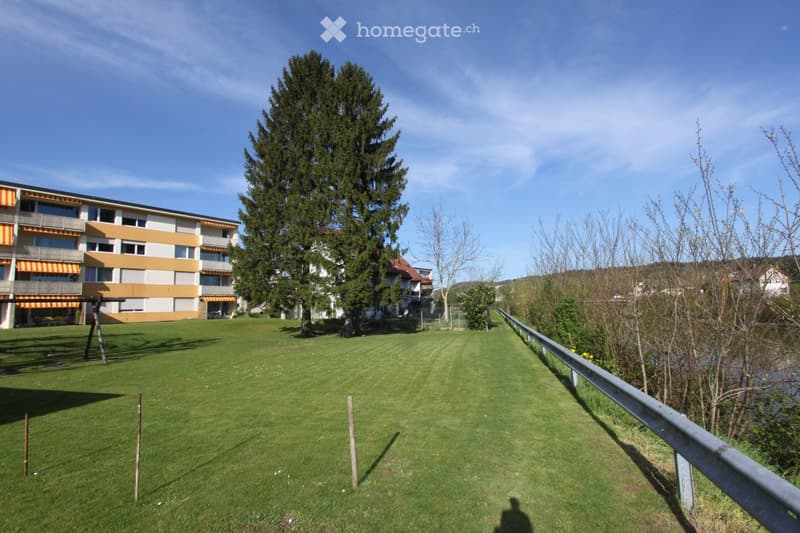 Well-maintained lawn area with a swing set, surrounded by trees and a multi-story building with balconies and orange accents.