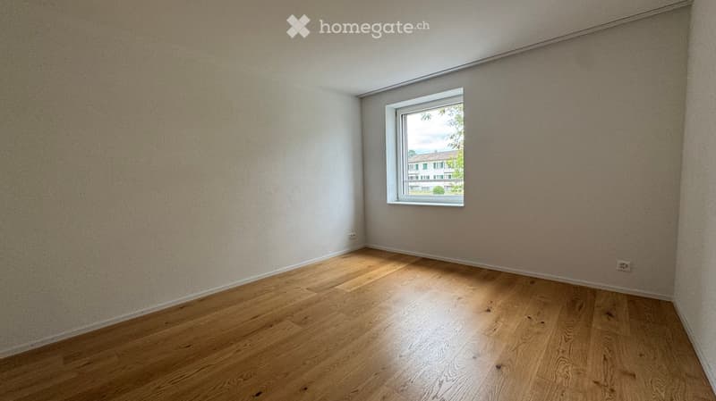 Empty room with wooden flooring and a single window showing a view of a neighboring building. The walls are painted in a neutral tone, and there are no furnishings or decorations present.