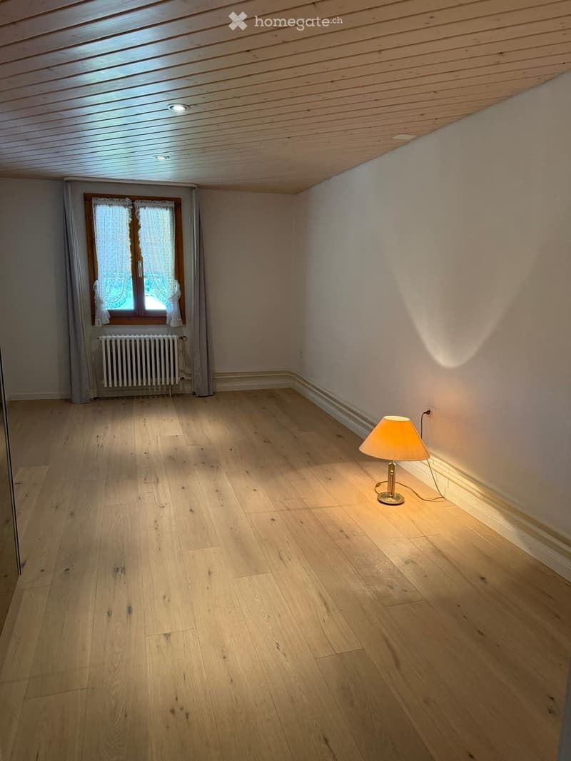 Empty room with wooden flooring, white walls, a window with curtains, a radiator, and a single table lamp.