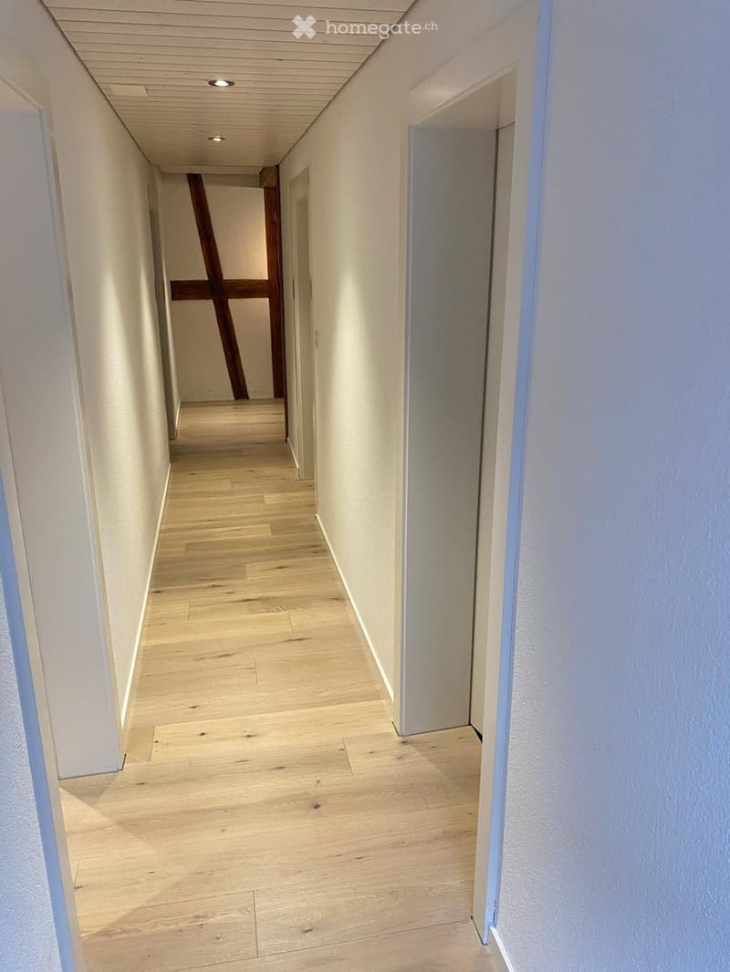 A narrow hallway with light wooden flooring, white walls, and recessed ceiling lighting. Features a glass door on the right and a wooden door on the left.