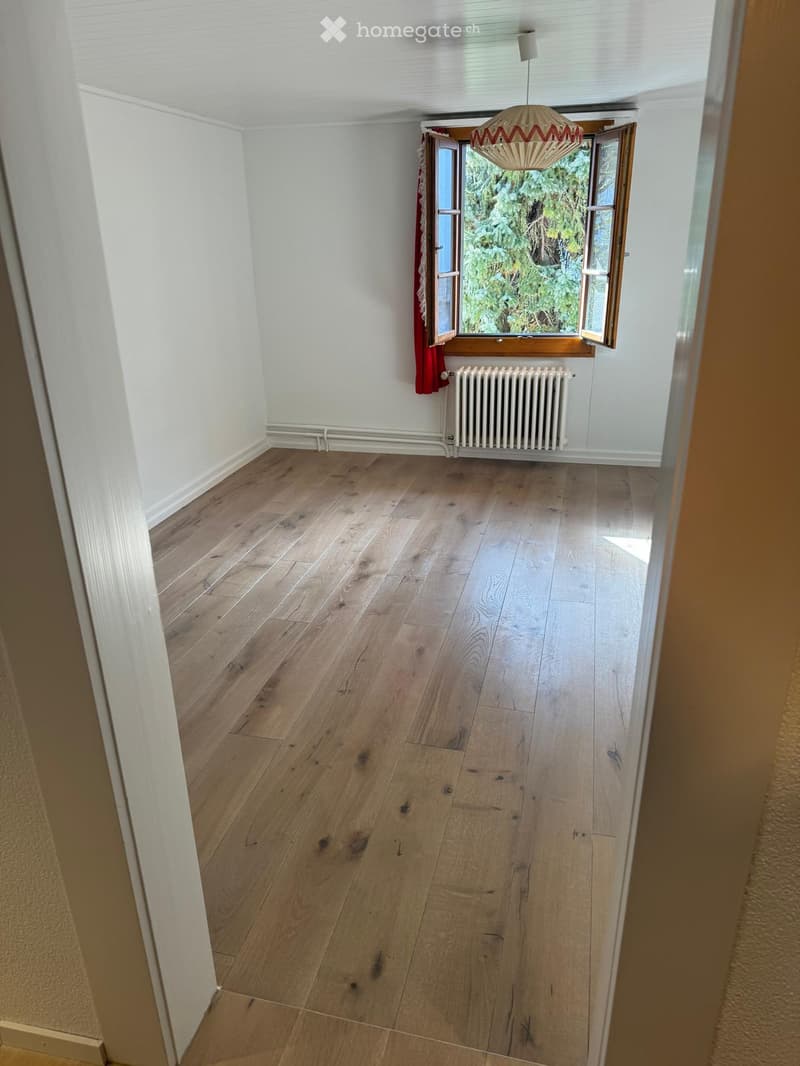 Empty room with wooden flooring, white walls, a radiator, and a window with red curtains. The room is well-lit with natural light coming through the window.