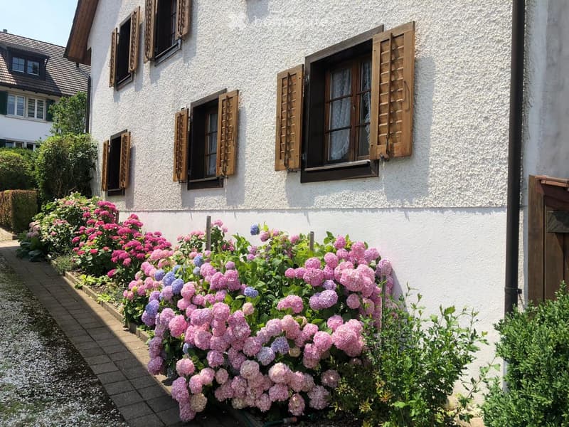 Two-story house with white exterior, wooden shutters, and a well-maintained garden with pink and purple flowers.