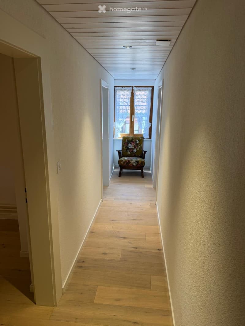 A long, narrow hallway with wooden flooring and light-colored walls. A single chair with a floral-patterned seat is placed against the far wall near a window with white curtains.
