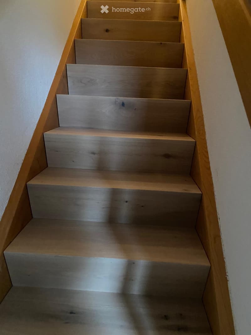 Wooden staircase with light-colored steps and handrails, set against white walls.