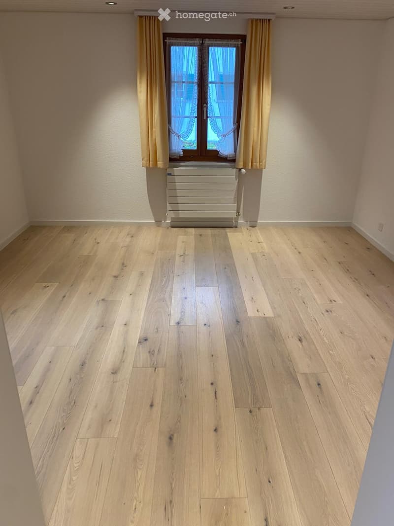 Empty room with light wooden flooring, white walls, and a window with yellow curtains and a radiator below it.