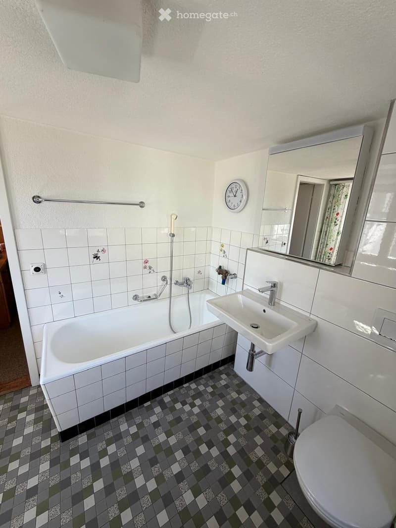 Bathroom with white tiled walls, a bathtub, a sink, a toilet, a mirror, and a clock on the wall.