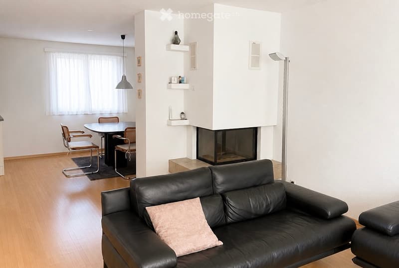 Modern living room with a black leather sofa, a small dining area with chairs and a table, a fireplace, and decorative shelves.