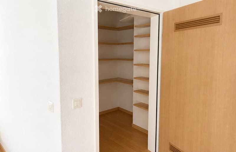 Empty closet with wooden shelves and a wooden door, featuring light-colored walls and wooden flooring.
