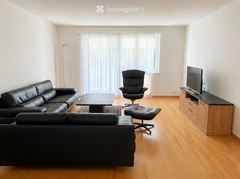 Modern living room with light wooden flooring, large window with white curtains, black leather sofa, coffee table, black chair, ottoman, and TV stand with flat-screen TV.