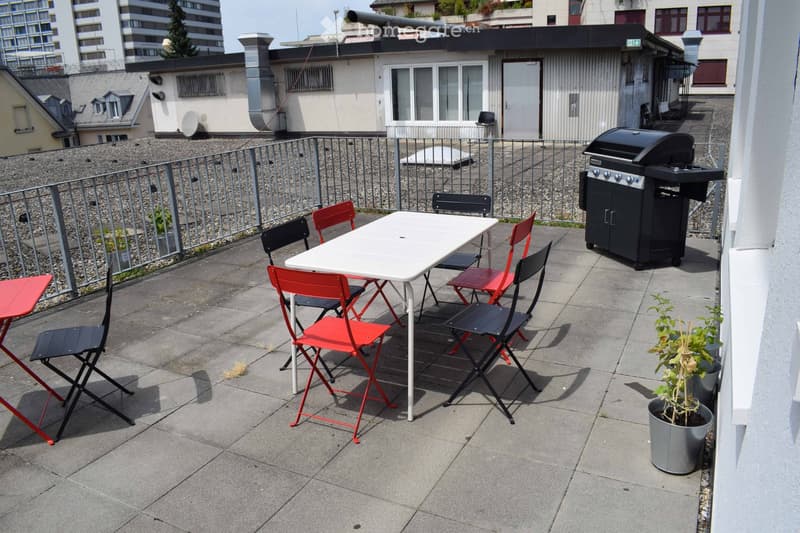 Roof terrace with a white table, red and black folding chairs, a black grill, and potted plants.