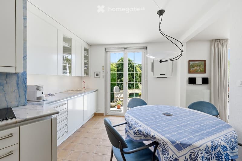 Bright kitchen with white cabinets, a dining table with blue chairs, and a coffee maker on the counter. Large sliding glass doors open to a patio with outdoor furniture.