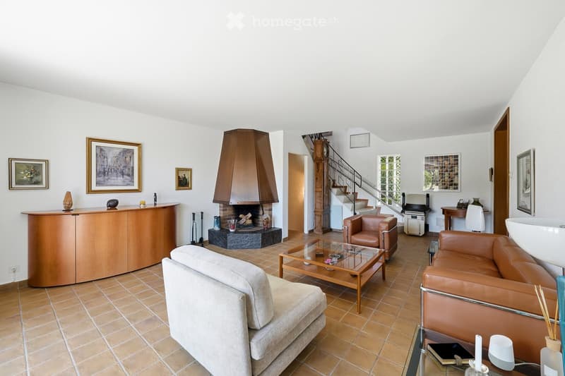 Spacious living room with a fireplace, leather armchairs, a sofa, a coffee table, and a bar cabinet. The room features tiled flooring and several framed pictures on the walls.