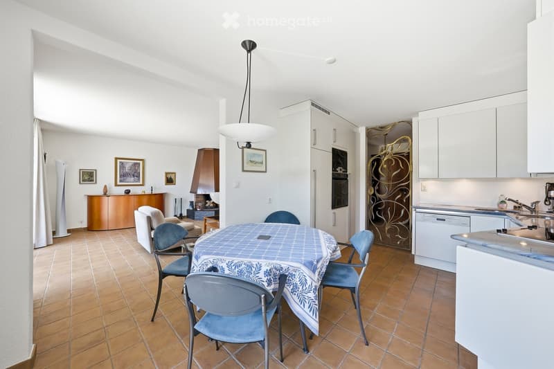 A well-lit, open-plan living and dining area with a tiled floor, a central dining table with chairs, a sofa, a fireplace, and a modern kitchen with white cabinets and stainless steel appliances.