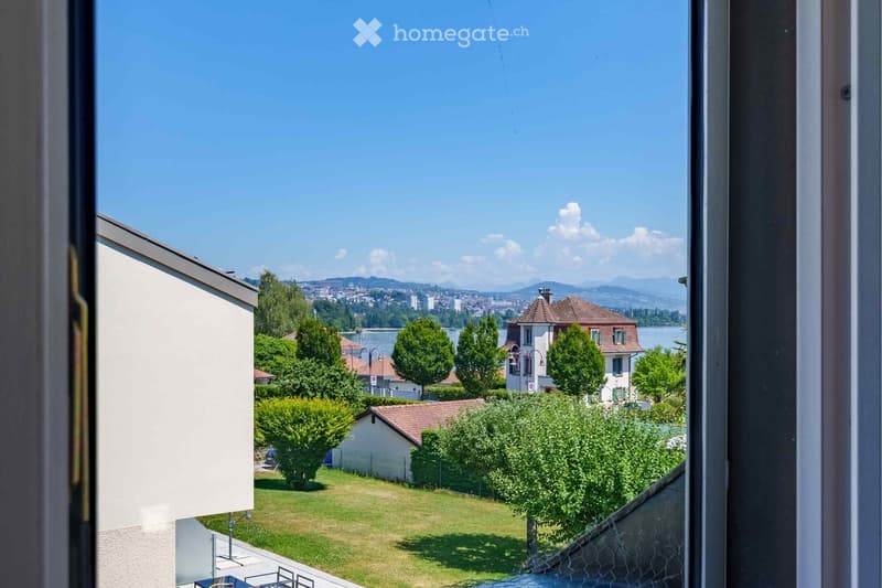 Balcony view overlooking a well-maintained garden with green grass, trees, and neighboring houses, with a distant view of a lake and cityscape under a clear blue sky.