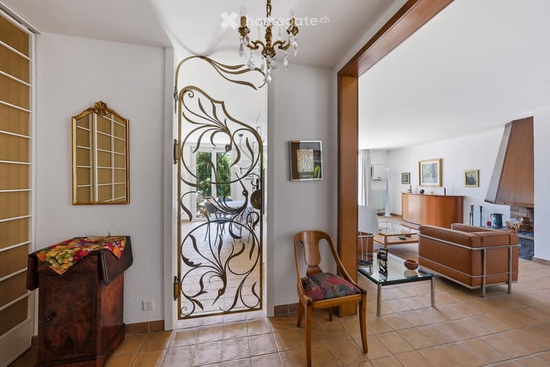 Elegant living room with a chandelier, sofa, coffee table, wooden chair, and a decorative cabinet. A large, ornate metal door leads to a dining area with additional furniture.