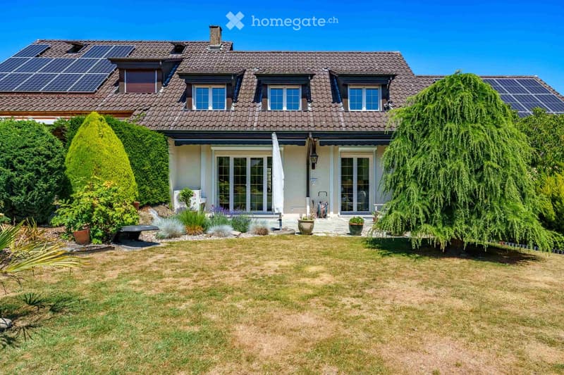 Two-story house with a tiled roof, solar panels, large windows, and a well-maintained garden with a lawn, shrubs, and potted plants.