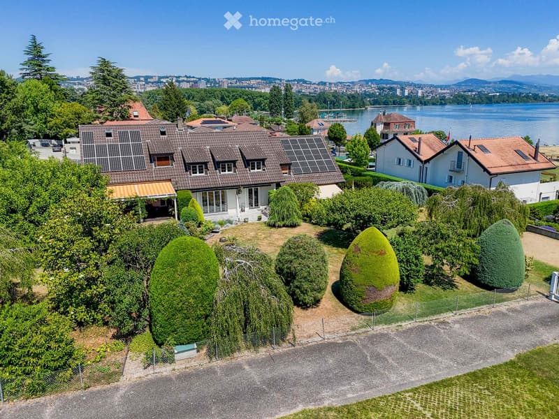 Aerial view of a two-story house with a tiled roof, surrounded by a well-maintained garden with trimmed hedges and trees, overlooking a lake and distant cityscape.