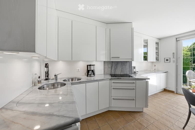 Modern kitchen with white cabinets, marble countertops, stainless steel sink, coffee maker, and a small seating area near the window.