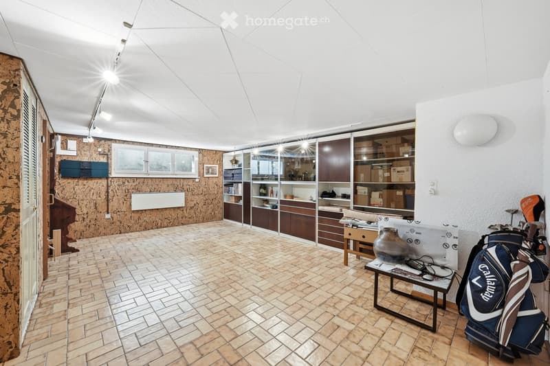 A spacious, well-lit room with a modern, curved ceiling and recessed lighting. The room features a mix of wooden and glass cabinetry, a small table with a vase, and a golf bag placed near the wall. The floor is tiled with a warm, earthy tone.
