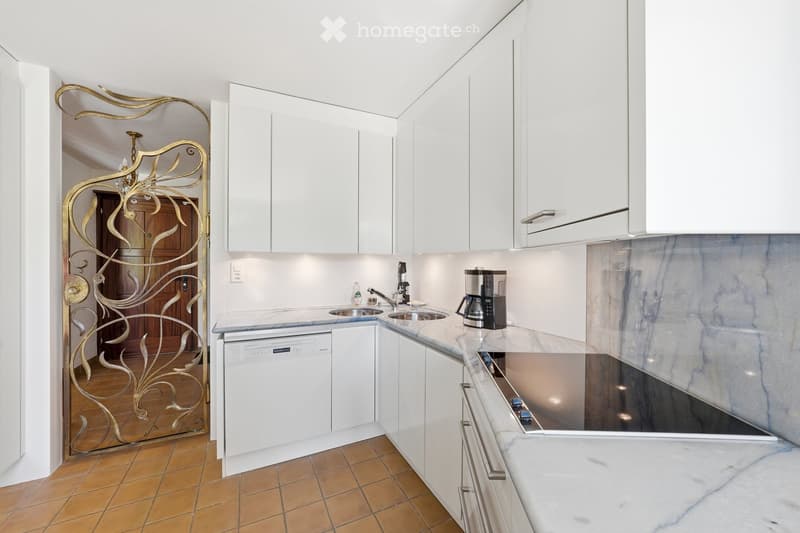 Modern kitchen with white cabinets, marble countertops, and stainless steel appliances, including a sink, faucet, coffee maker, and dishwasher. The floor is tiled in warm tones, and a decorative golden door is visible in the background.