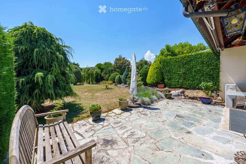A well-maintained garden with a stone-paved patio, wooden bench, potted plants, and a clear view of a grassy lawn and trees.