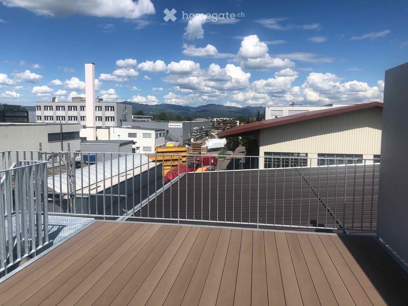 Wooden deck with metal railing, overlooking an industrial area with buildings and a distant mountain range under a partly cloudy sky.