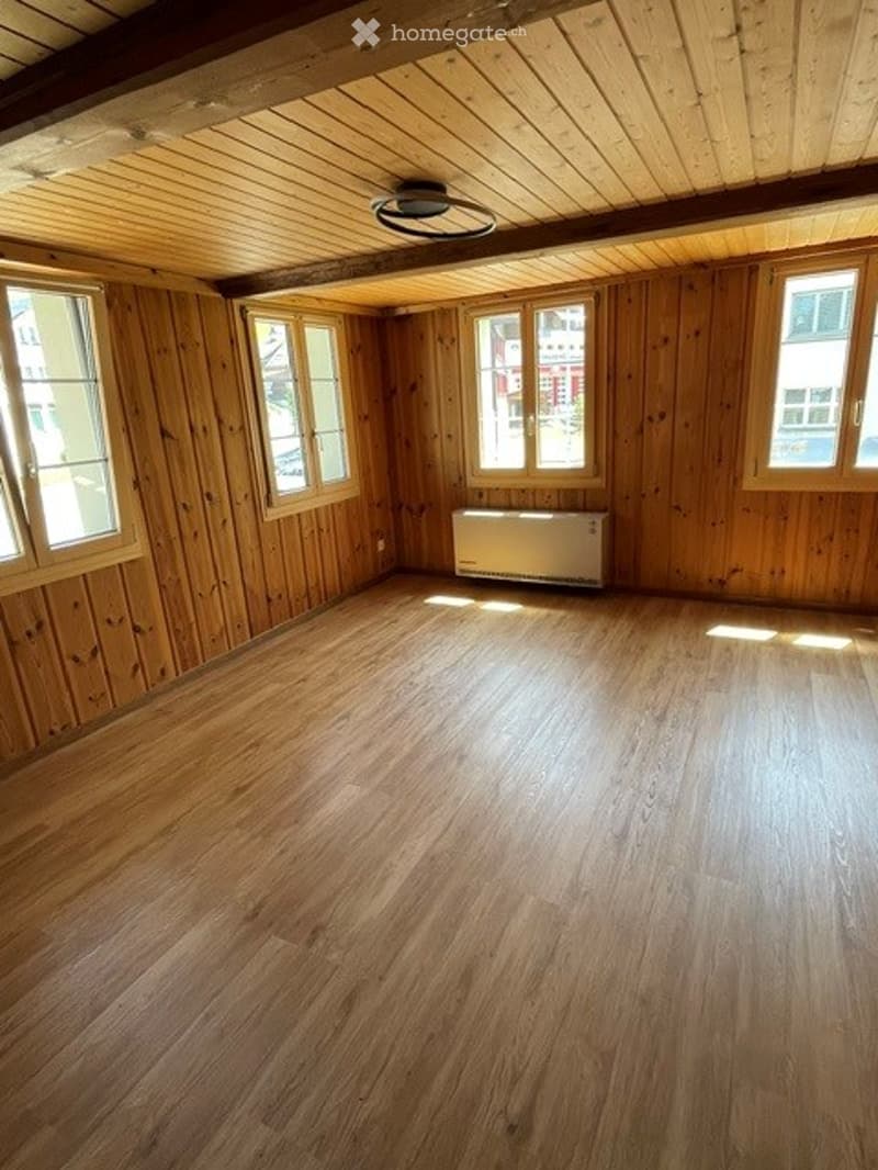 Empty room with wooden walls and floor, large windows, and a radiator. No furniture or equipment.
