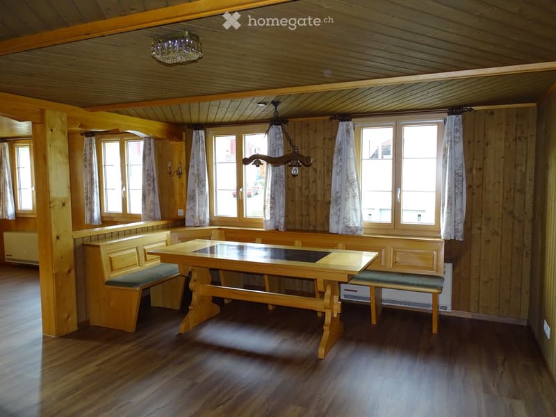Wooden kitchen with a central table, benches, and large windows with curtains. Exposed wooden beams on the ceiling and wooden walls.