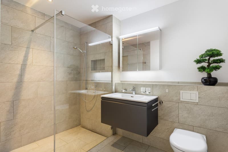 Modern bathroom with a large mirror, double sink, toilet, and a walk-in shower with glass enclosure. Beige tiles and a sleek black cabinet.