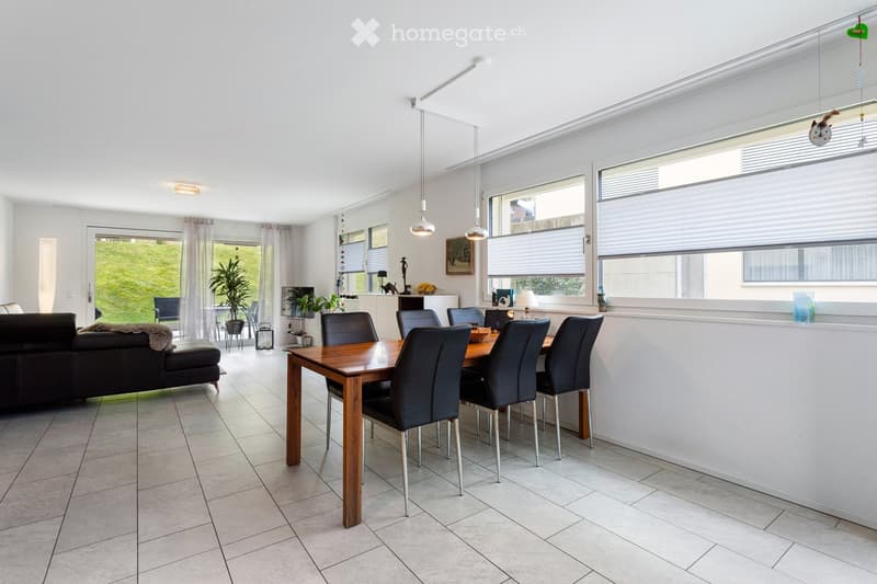 Bright, open living and dining area with a dark leather sofa, wooden dining table with six chairs, pendant lights, and large windows with blinds.