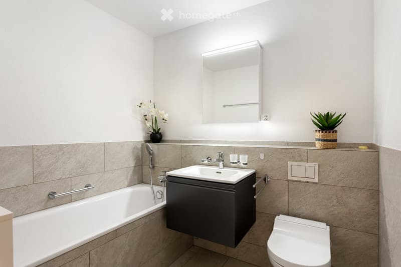 Modern bathroom with a bathtub, sink, toilet, and a large mirror above the sink. The walls are tiled in a neutral color, and there are decorative plants on the counter.