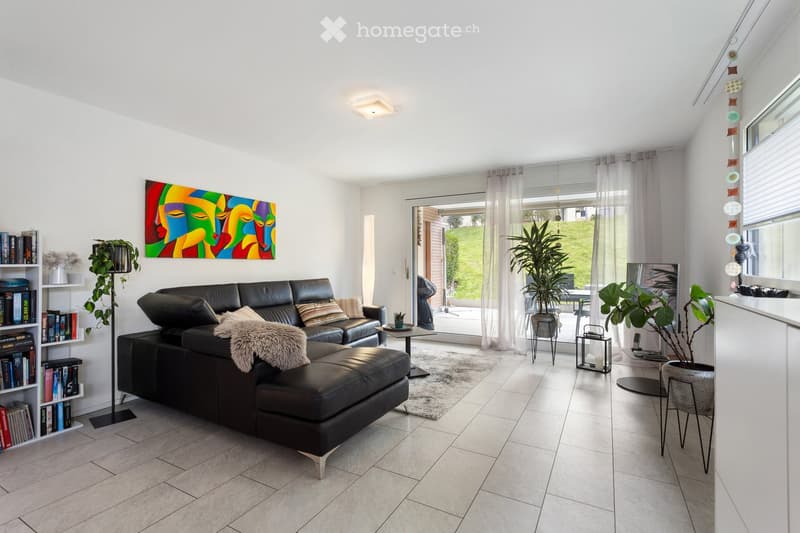 Modern living room with a black leather sectional sofa, throw blanket, coffee table, bookshelf, floor lamp, and large windows leading to a patio.
