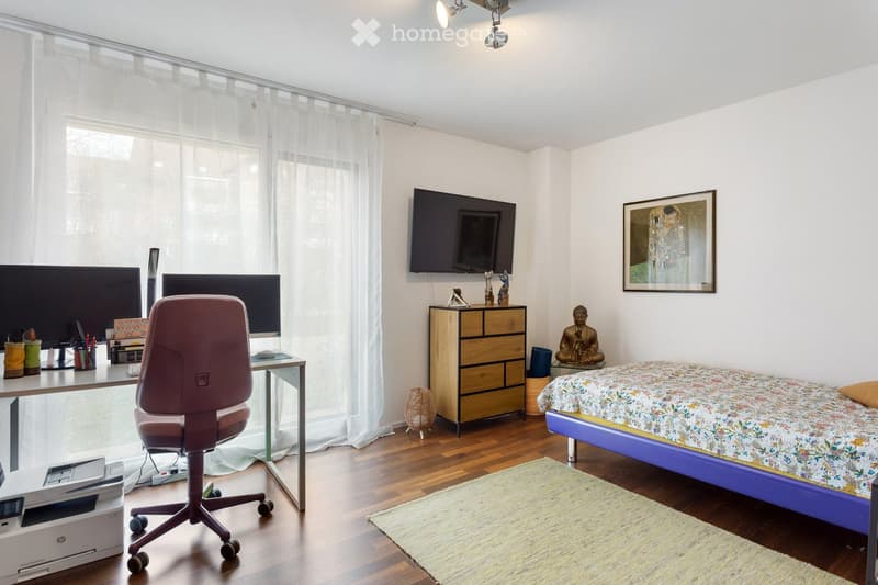 A well-lit bedroom with a bed, desk, chair, and a TV mounted on the wall. The room features a wooden floor, large windows with sheer curtains, and decorative items including a picture frame and a small statue.