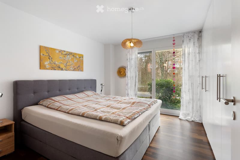 Modern bedroom with a grey upholstered headboard, a plaid bedspread, a wooden nightstand, a large window with sheer curtains, and a decorative wall piece.