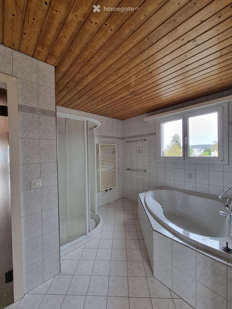 modern and spacious bathroom with a bathtub and shower stall, white tiled walls and floor, wooden ceiling, and window with a view of the outside.
