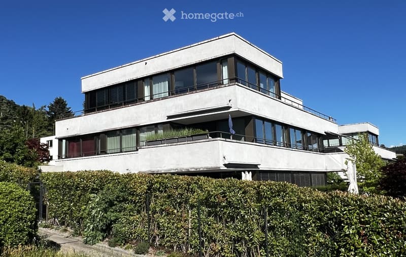 Modern two-story house with white facade, large windows, balconies, and surrounding greenery.