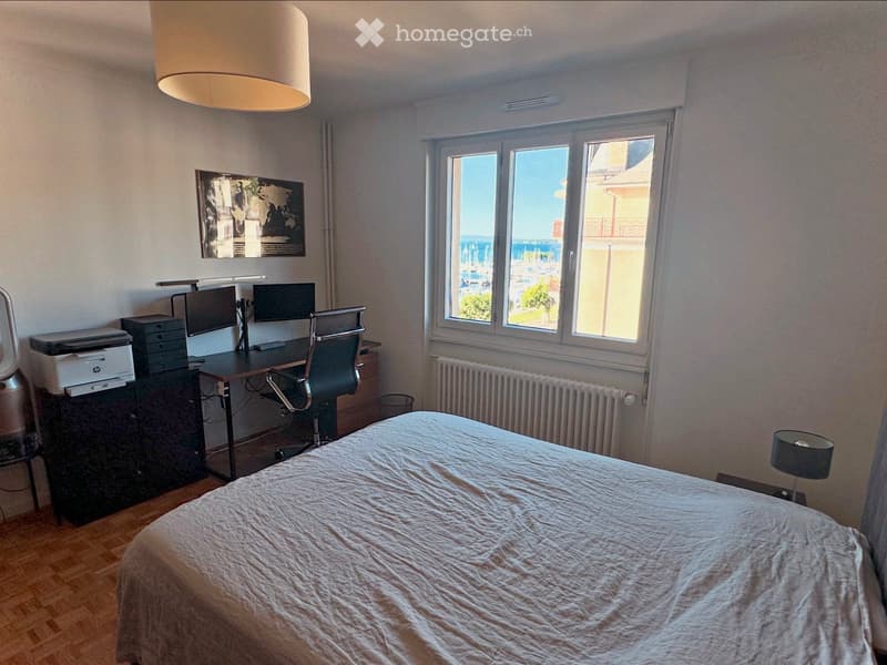 Well-lit bedroom with a large bed, desk with a computer and chair, and a view of the sea through large windows.