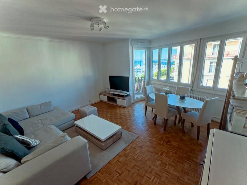 Open-plan living and dining area with wooden flooring, large windows offering a view, a grey sofa with cushions, a coffee table, an ottoman, a dining table with chairs, and a television on a stand.