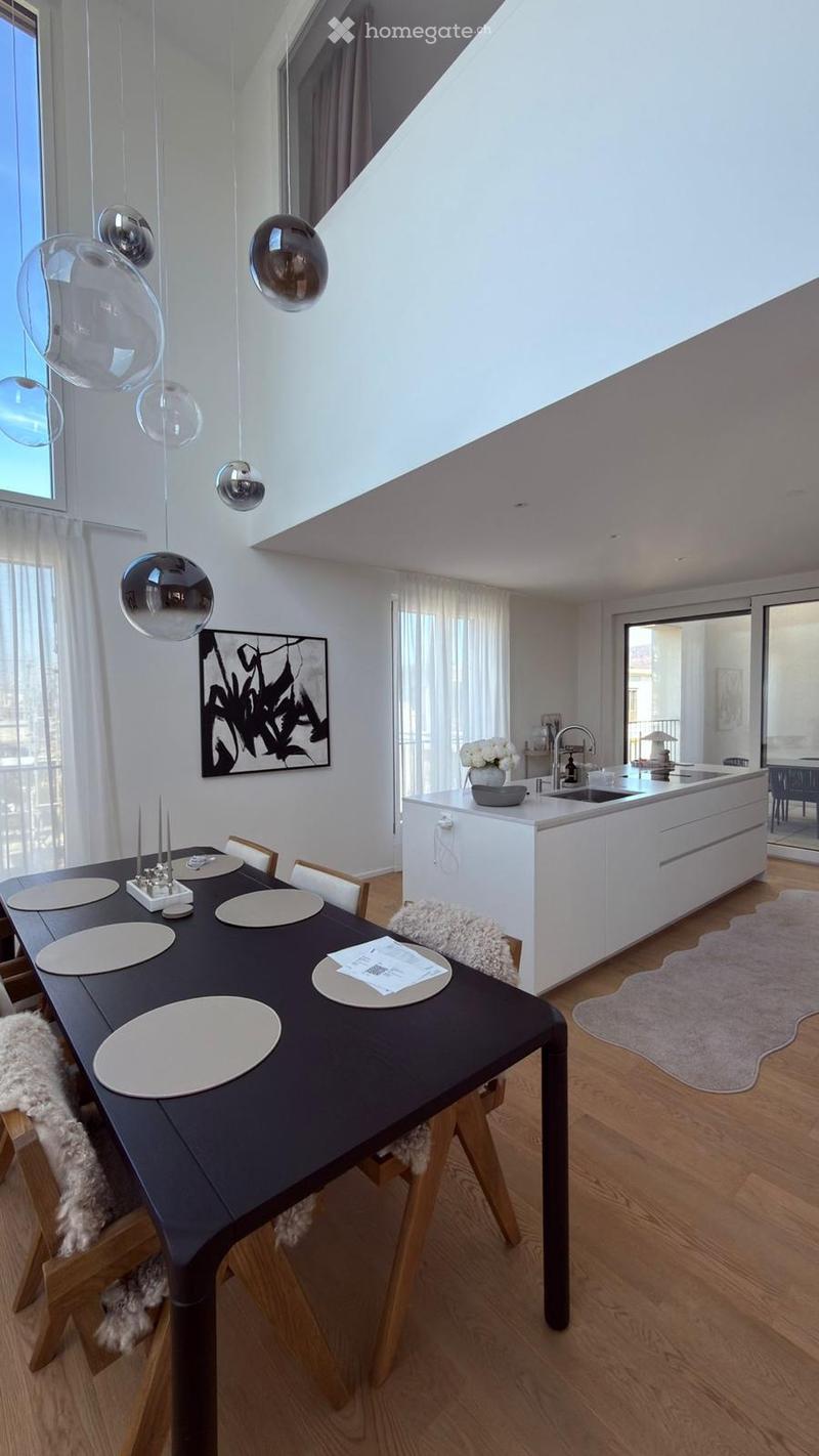 Modern dining area with a dark wooden dining table, white chairs, pendant lights, and a white kitchen island with a sink and faucet. A vase with white flowers sits on the island, and a picture frame hangs on the wall.