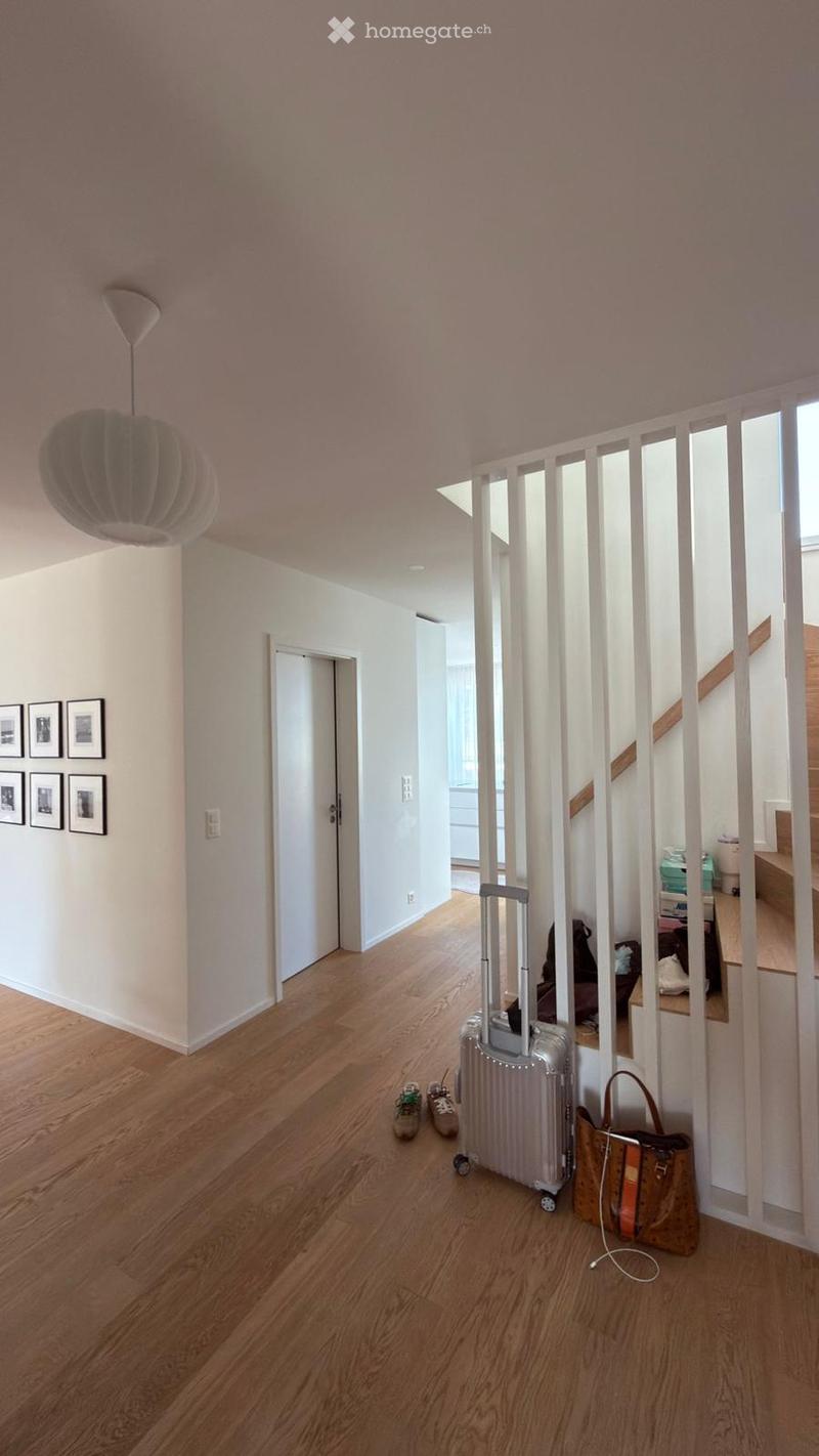 A modern hallway with light wooden flooring, white walls, and a staircase leading upwards. A large, round pendant light hangs from the ceiling, and a set of framed pictures is displayed on one wall. A silver suitcase, a pair of sneakers, and a brown handb