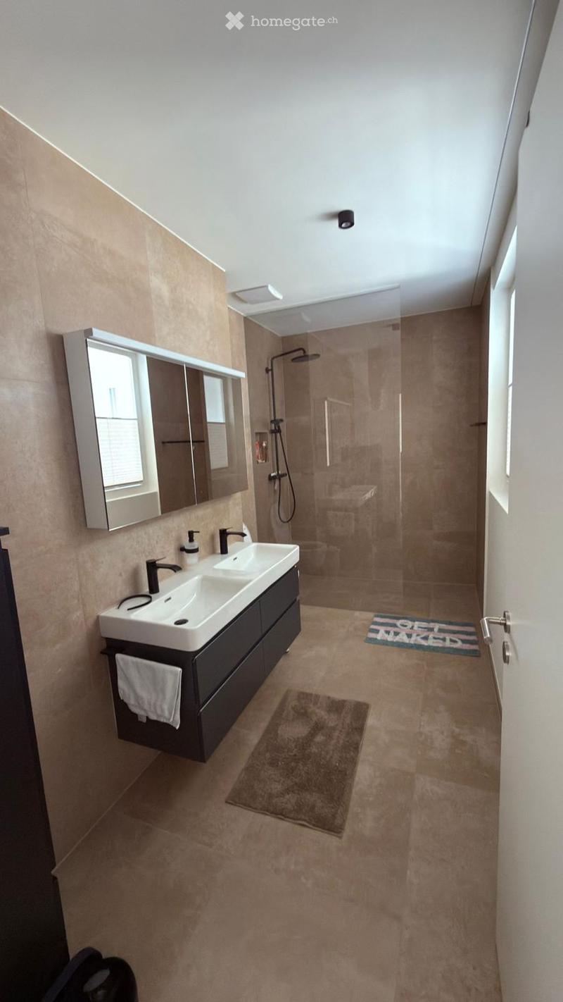 Modern bathroom with double sink, large mirror, glass-enclosed shower, beige tiles, and a small rug.