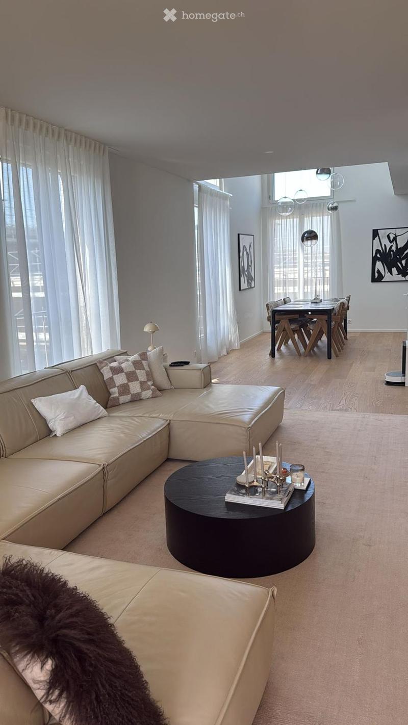 Modern living room with a beige sectional sofa, round coffee table, dining table with chairs, and large windows with white curtains.
