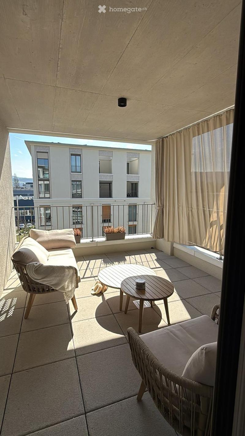 Balcony with modern furniture including a sofa, small round table, and wicker chairs, with a view of neighboring buildings.