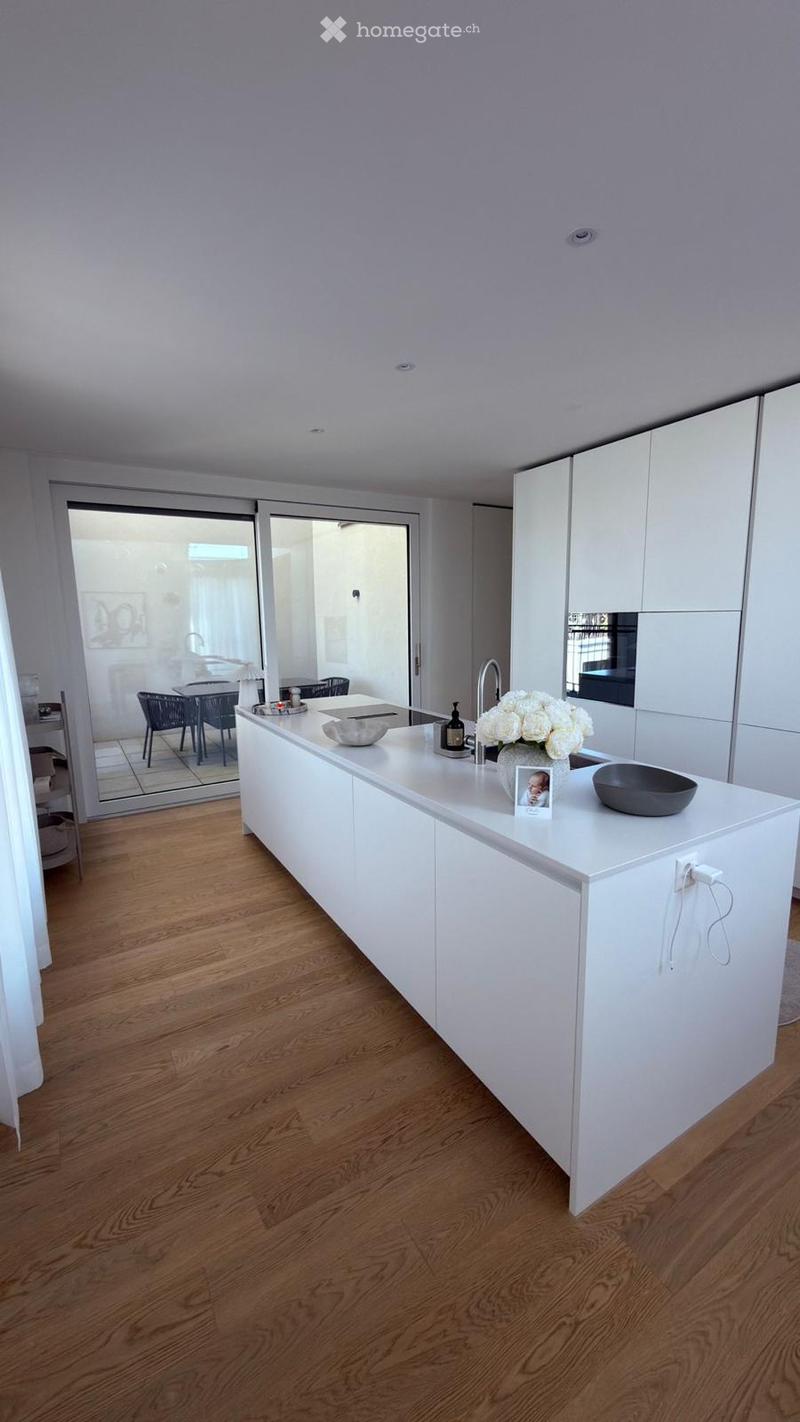 Modern kitchen with white cabinetry, a central island, and a wooden floor. The island features a sink, a faucet, a bowl, a flower arrangement, and a picture frame. A dining area with chairs and a table is visible through a large glass door.